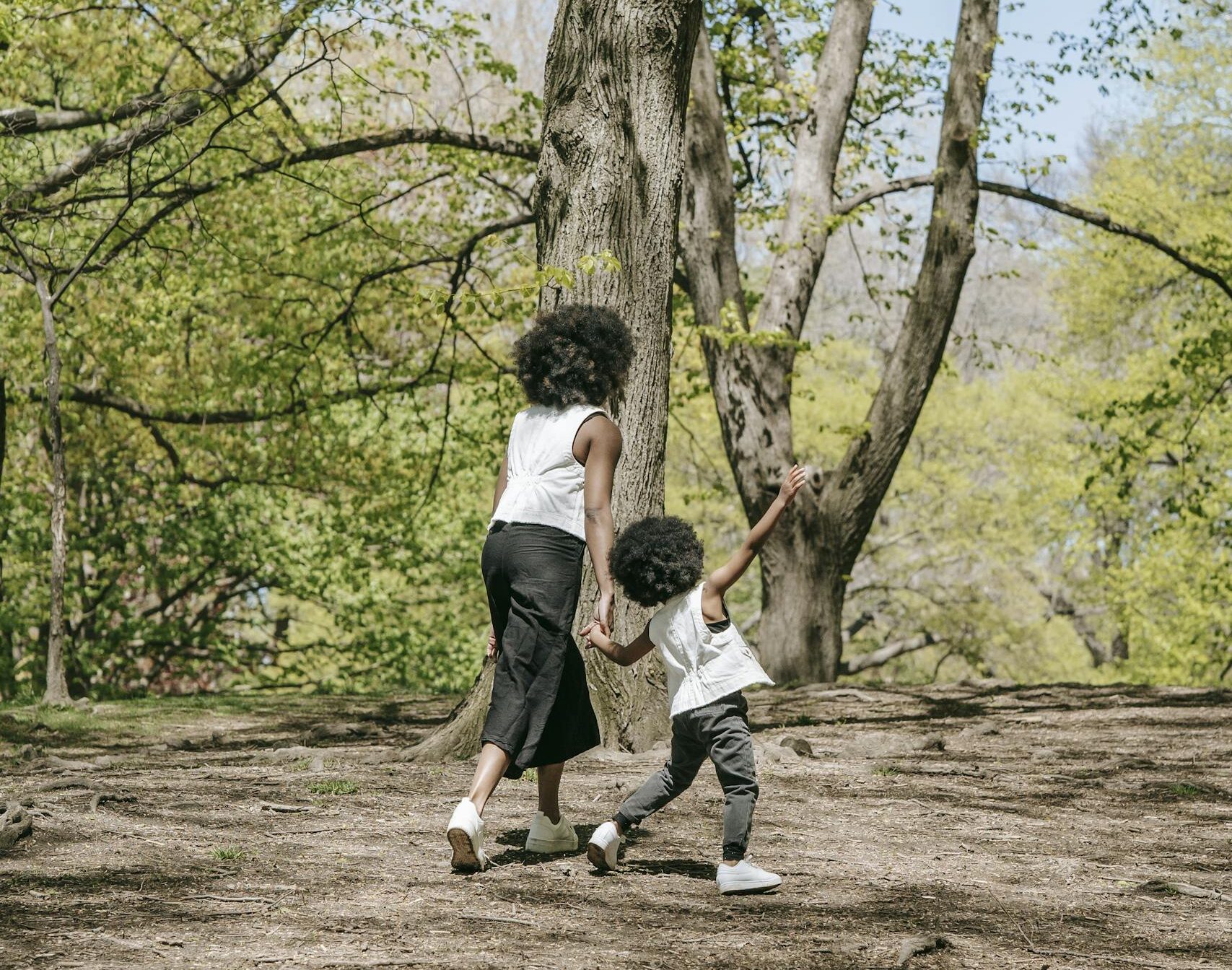 A mother and son walk hand-in-hand through a lush, sunny forest in spring.